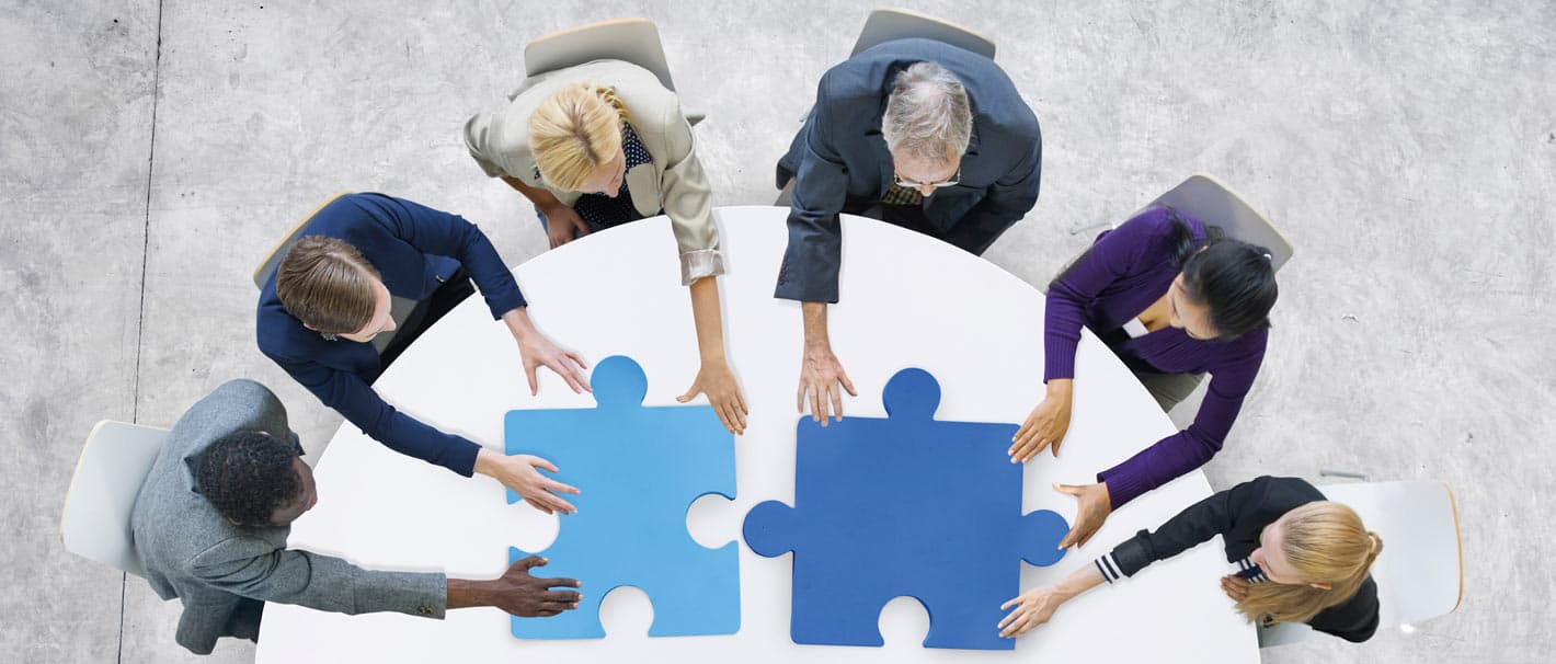Sechs Menschen, die gemeinsam um einen Tisch herum sitzen, fügen zwei Teile eines Puzzles zusammen.