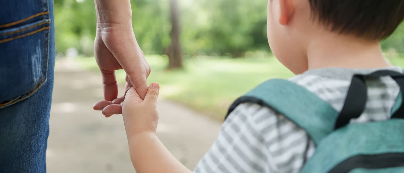 Aufnahme von hinten: Eine Mutter und ihr kleiner Sohn laufen Hand in Hand. Der Sohn trägt einen Rucksack.