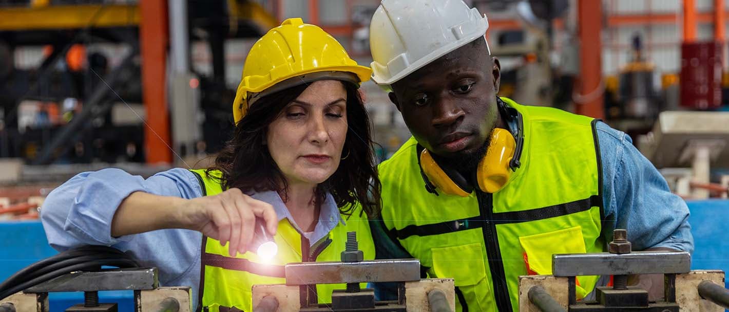 Eine Frau und ein Mann stehen in einer Fabrikhalle und schauen gemeinsam in eine Maschine, die die Frau mit einer Taschenlampe beleuchtet. Beide tragen Schutzhelme und Warnwesten.