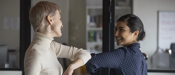 Zwei Frauen berühren sich gegenseitig mit ihren Ellbogen und lächeln sich an.
