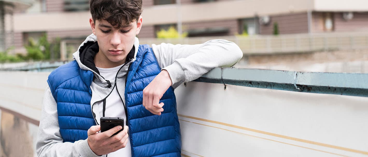 Ein Jugendlicher steht vor einem Wohnblock und schaut auf sein Handy, das er in der Hand hält.