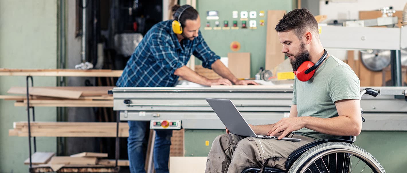 Ein Mann im Rollstuhl arbeitet in einer Schreinerei an einem Laptop, den er auf dem Schoß hat. Im Hintergrund ist sein Kollege zu sehen.