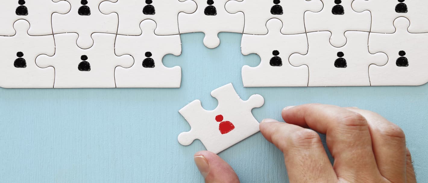 Eine Hand fügt ein Puzzlestück mit einer roten Figur in ein Puzzle mit vielen schwarzen Figuren ein.