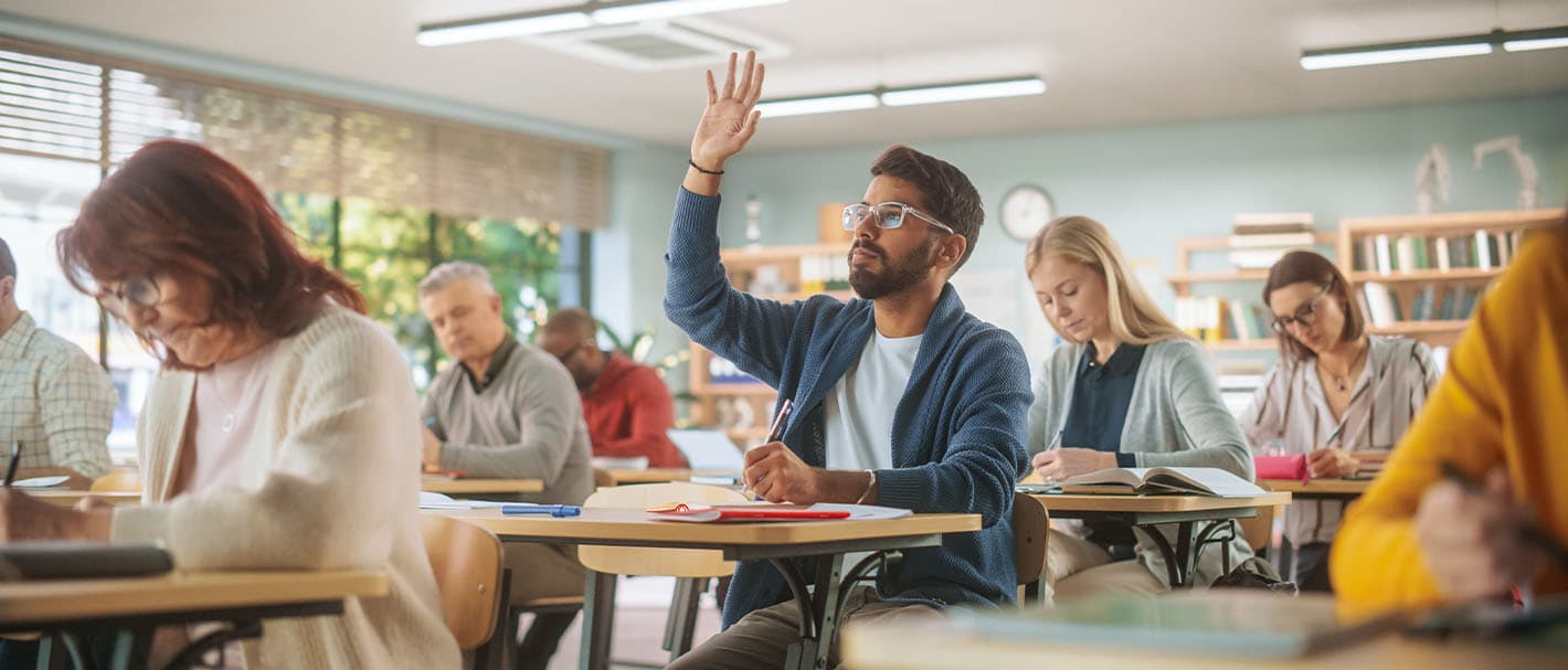 Frauen und Männer sitzen an Tischen, angeordnet wie in einer Schule. Ein Mann hebt seine Hand, um sich zu melden.