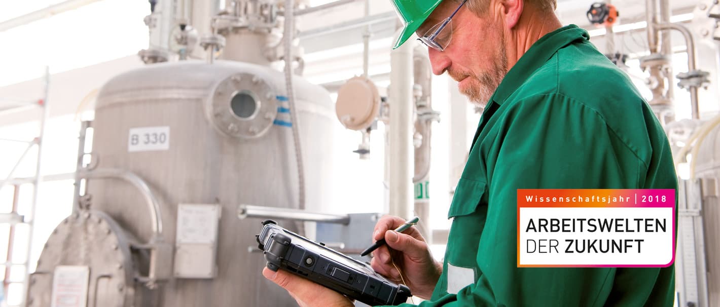 Älterer Herr mit grünem Arbeitsanzug hält elektronischen Stift über einem Tablet vor einem großen Kessel in einer Fabrikhalle.