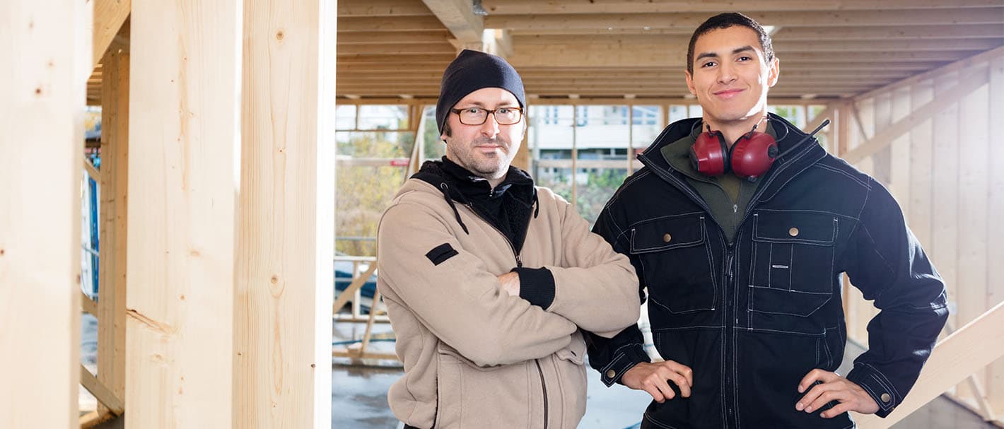 Portrait von zwei Zimmerern, die selbstbewusst auf ihrer Baustelle stehen und zum Betrachter blicken.