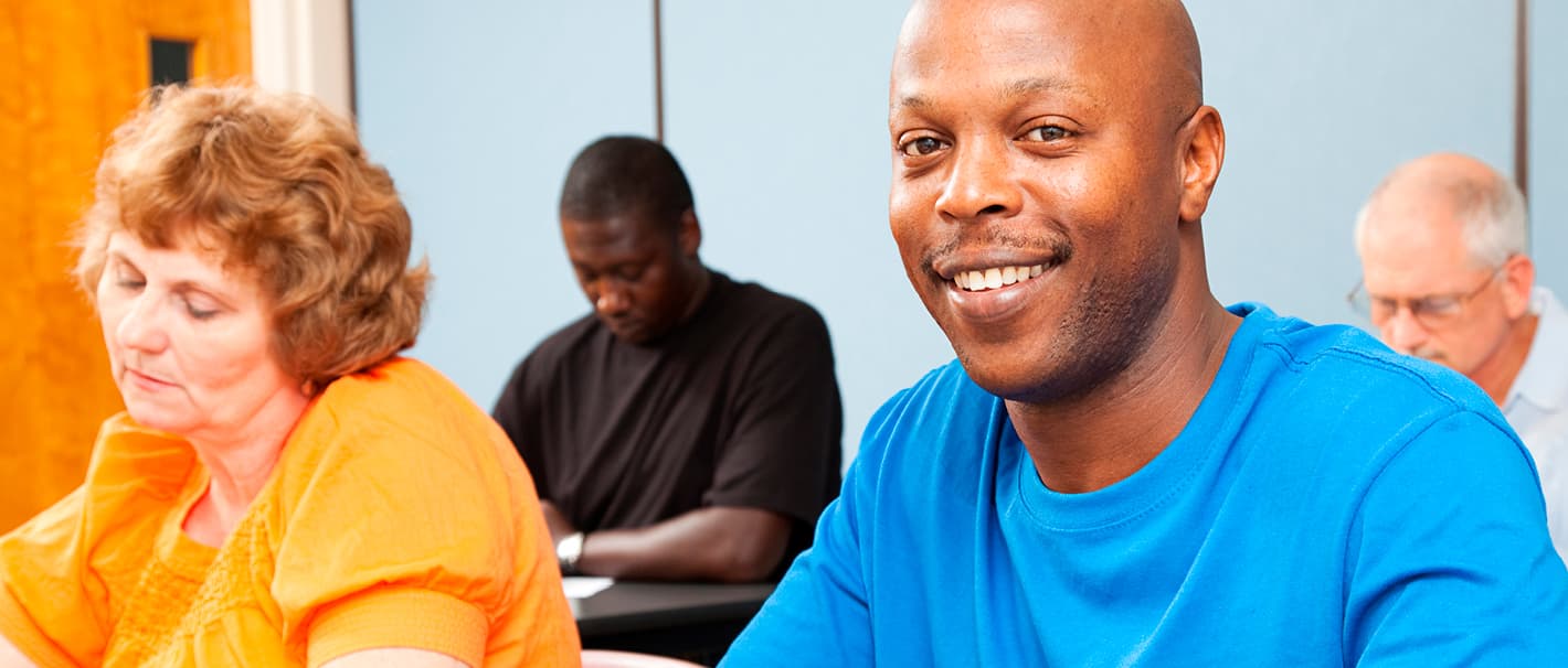 Mann mit dunkler Hautfarbe sitzt zusammen mit anderen Personen in einem Klassenzimmer