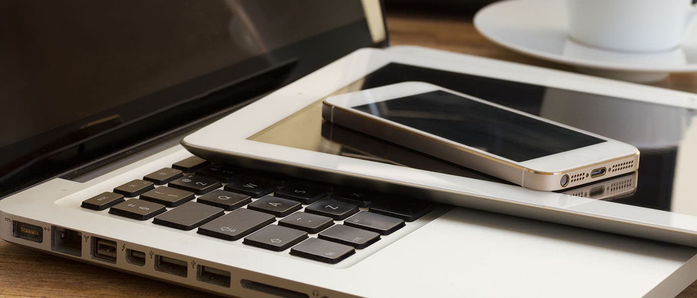 Auf einem Holztisch liegen gestapelt unten ein Laptop, darauf ein Tablet und oben ein Smartphone.
