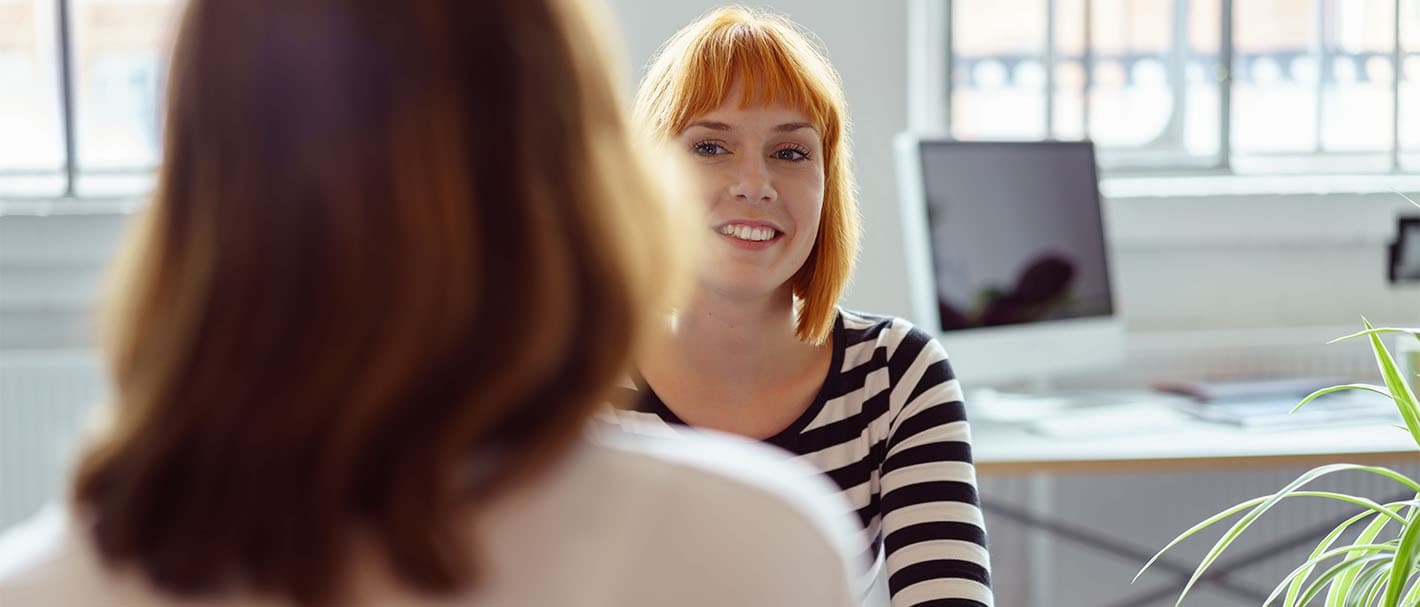 Zwei Frauen sitzen sich in einem Büro bei einem Gespräch gegenüber. Die eine Frau ist dem Betrachter zugewandt, die andere ist von hinten abgebildet.
