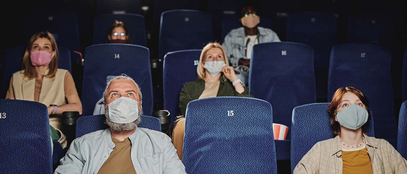 Frontalaufnahme von mehreren Männern und Frauen, die sich einen Film im Kino ansehen. Sie tragen alle einen Mundschutz und zwischen ihnen ist jeweils ein Sitz frei.
