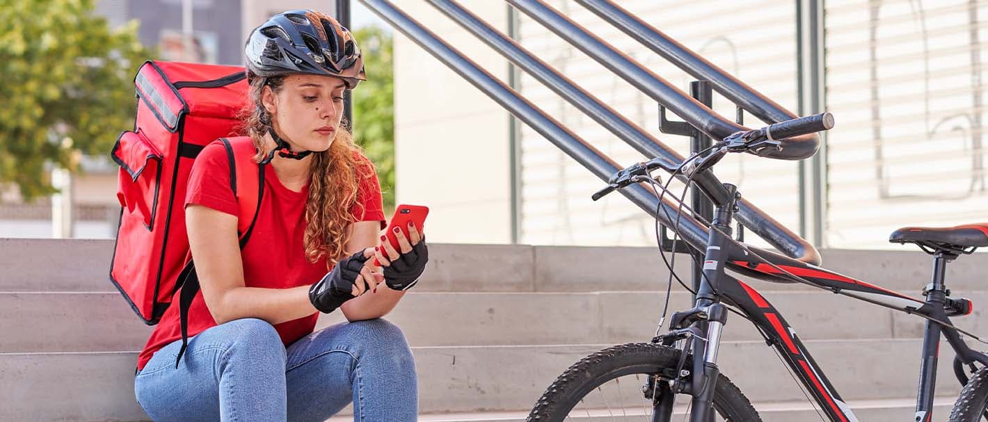 Fahrradkurierin sitzt neben ihrem Fahrrad auf einer Treppe und überprüft auf ihrem Handy, ob neue Aufträge vorliegen.