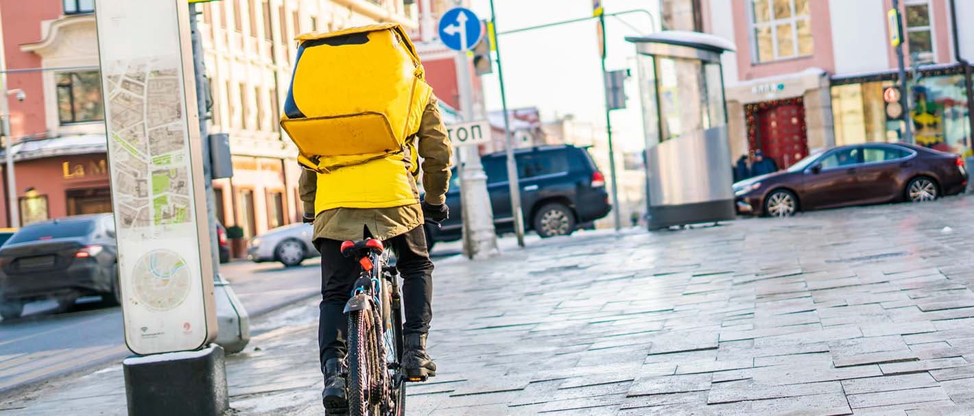 Rear view of a male bicycle courier carrying a yellow delivery bag on his back.