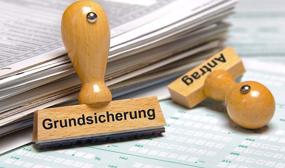Zwei Stempel liegen vor einem Stapel mit Unterlagen. Einer Trägt sie Aufschrift „Grundsicherungsforschung“ einer das Wort „Antrag“.