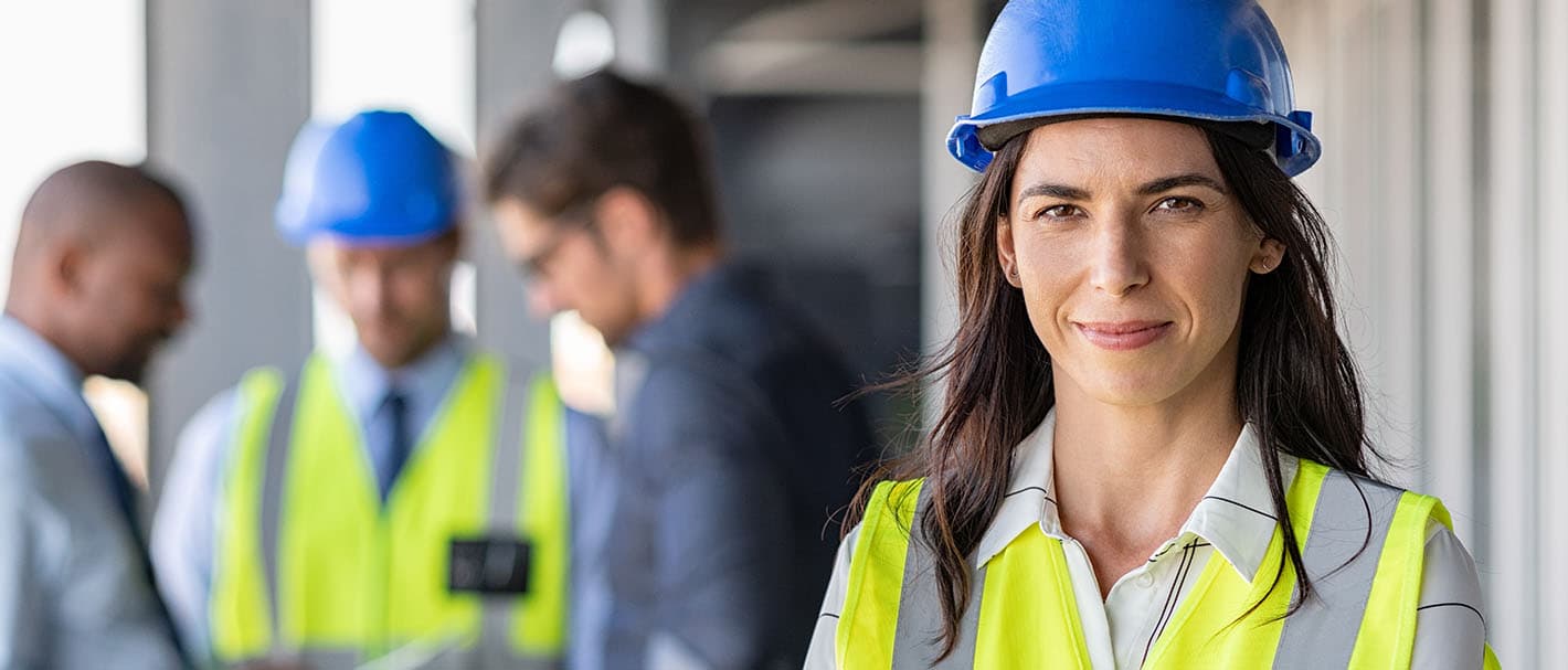 Portrait of a female architect standing on a construction site, smiling confidently. In the background, a group of men are looking at a plan. The focus is on the woman.