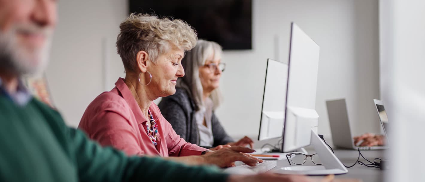 Perspektivische Aufnahme von erwachsenen Kursteilnehmern, die nebeneinander in einer Reihe an Computern arbeiten. Im Fokus ist eine Frau, die in der Mitte sitzt.