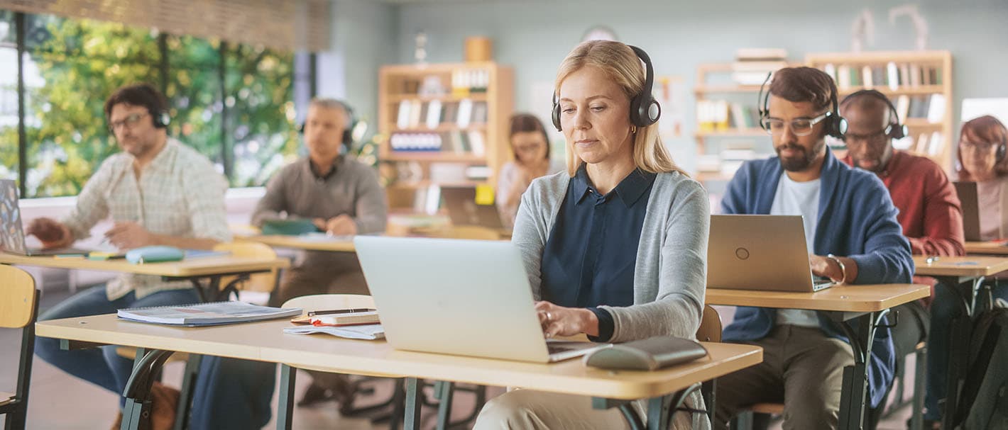 Adult men and women of different ages each sit at a desk in a training room and work with headsets on their laptops.
