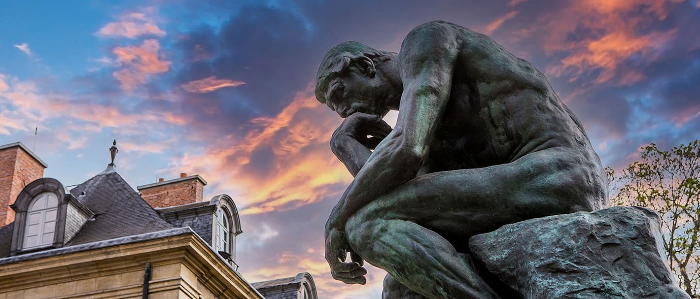 Das Foto zeigt Augustin Rodins Plastik “Der Denker” im Garten des Musée Rodin in Paris. Das Foto wurde seitlich und aus der Froschperspektive aufgenommen, vor einem Himmel mit orangenen und graublauen Wolken und einem Gebäude.
